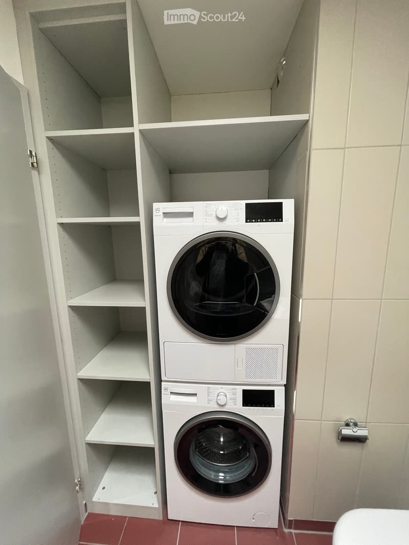 A compact bathroom with a stacked washing machine and dryer unit, white shelves on the left, and red tiled flooring.