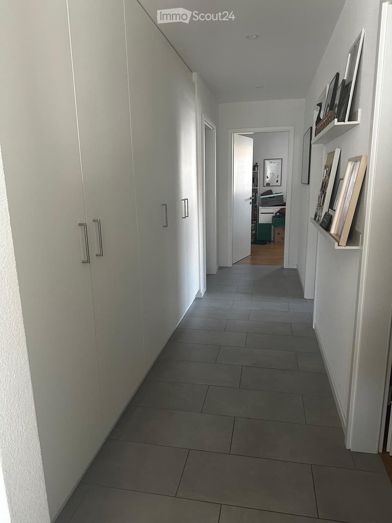 A clean, modern hallway with tiled flooring, white walls, and built-in shelving on one side. The hallway leads to an open door at the end, revealing a glimpse of another room with wooden flooring.