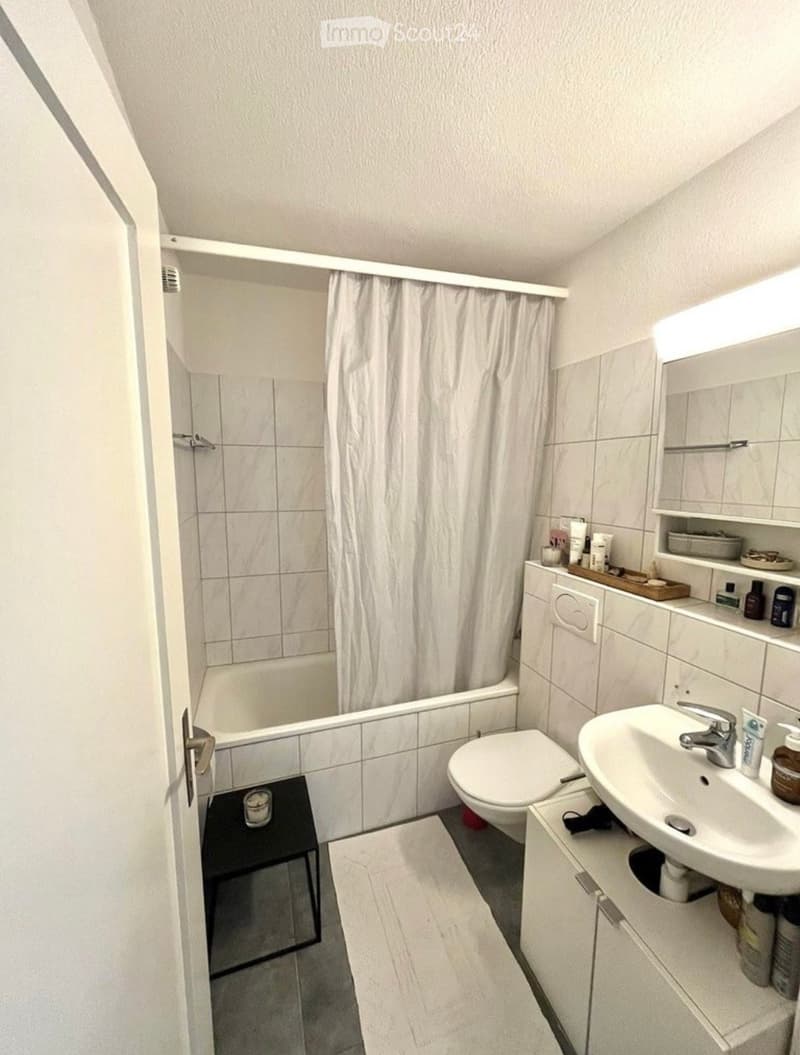Modern bathroom with a white sink, toilet, bathtub, and shower curtain. Includes a mirror, cabinet, and a small black stool near the door.