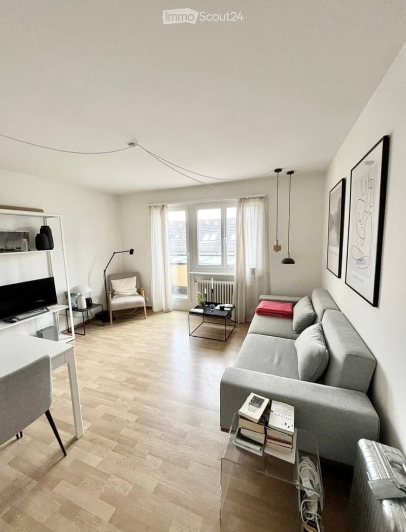 Modern living room with light wooden flooring, grey sofa, coffee table, armchair, and a TV on a stand. The room features large windows with white curtains and pendant lights hanging from the ceiling.