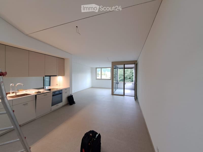 Modern kitchen with white cabinets, stainless steel sink, and built-in oven. Open space with large windows and sliding glass doors leading to a balcony.