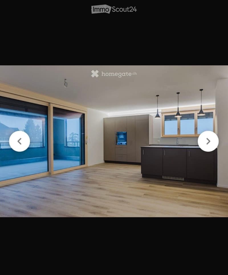 Modern kitchen with light wood flooring, grey cabinets, and a large window overlooking a balcony. The space is clean and well-lit with pendant lights above the counter.