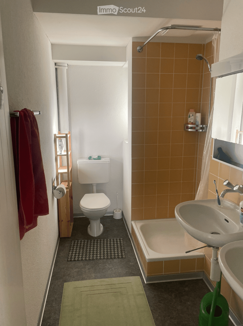 A compact bathroom with a toilet, bathtub, sink, and a small shelf. The walls are tiled in a warm orange color, and the floor is dark grey with a green bath mat. A towel hangs on the left side, and a toothbrush and toothpaste are visible on the sink.