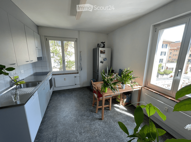 Modern kitchen with white cabinets, stainless steel sink, refrigerator, small wooden table with chairs, and potted plants. Large windows provide natural light.