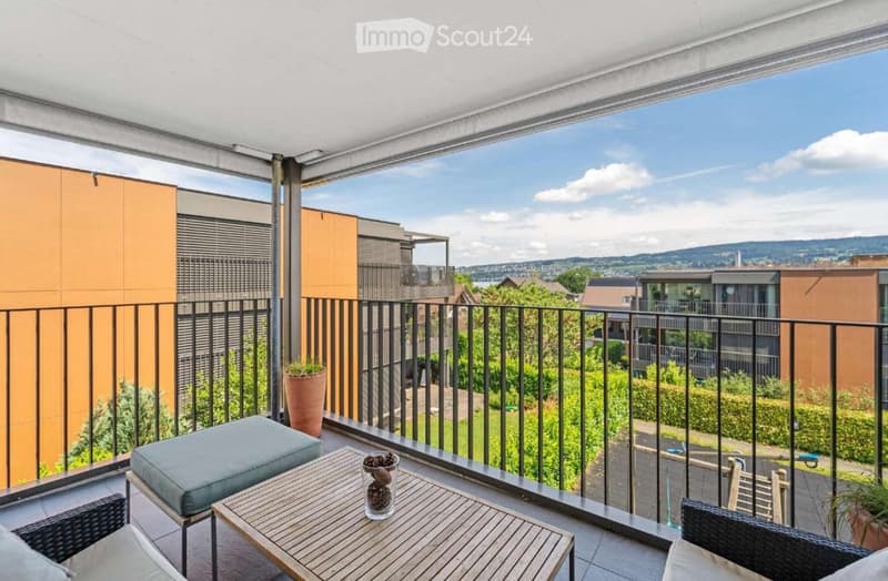 Balcony with wooden table, grey cushioned stool, wicker chairs, and potted plants, overlooking a green area with modern buildings.