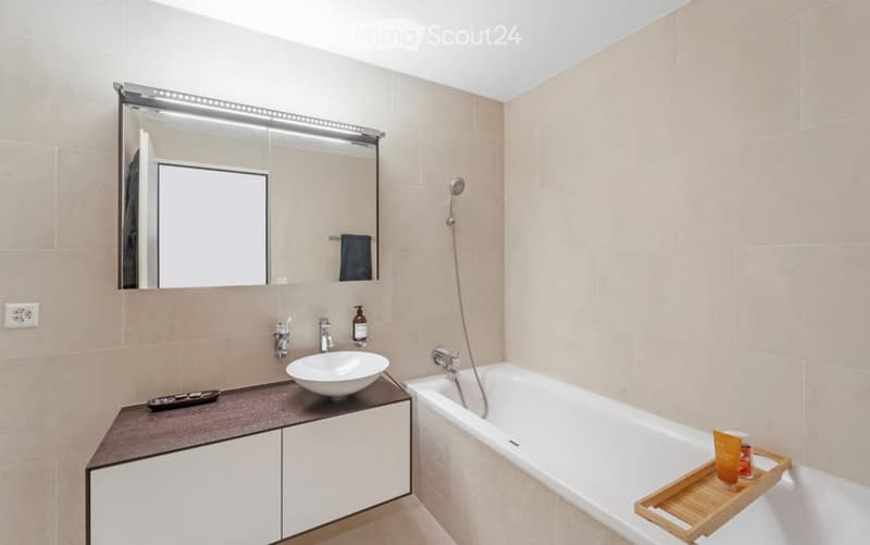 Modern bathroom with a white bathtub, a sink with a modern faucet, a large mirror with built-in lighting, and a towel rack. A small tray with toiletries is placed on the edge of the bathtub.