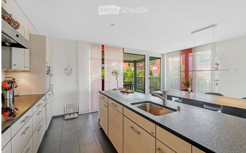 Modern kitchen with light wooden cabinets, granite countertop, stainless steel sink, and a small dining area with a table and chairs. The space features large windows with blinds and a view of a balcony.