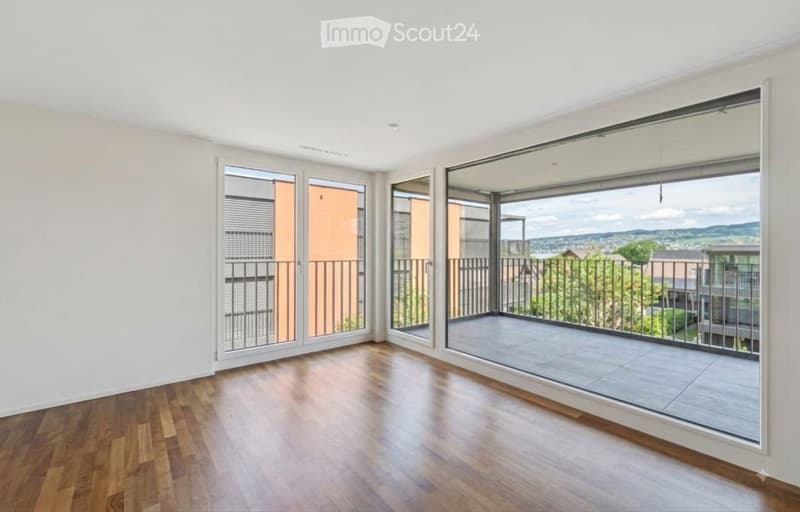 Empty room with wooden flooring, large glass sliding doors leading to a balcony with metal railings, and a scenic view of the surrounding area.