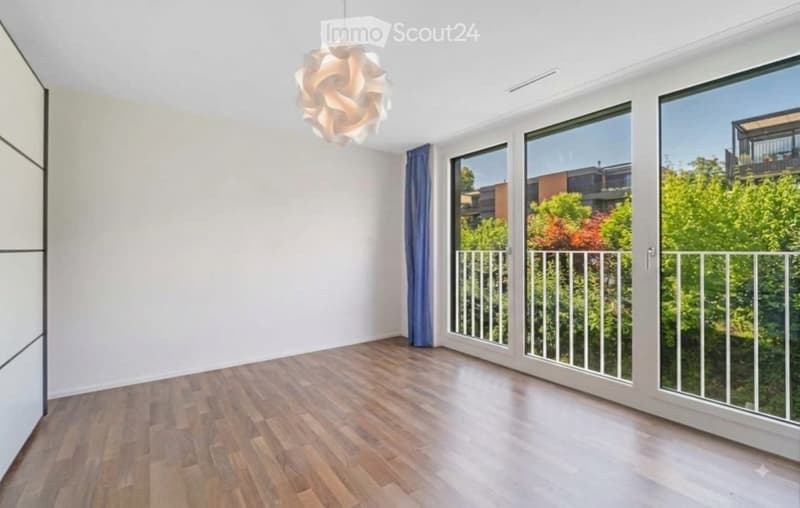 Empty room with wooden flooring, large windows, and a view of greenery outside. The room features a modern pendant light fixture and a sliding door leading to a balcony.