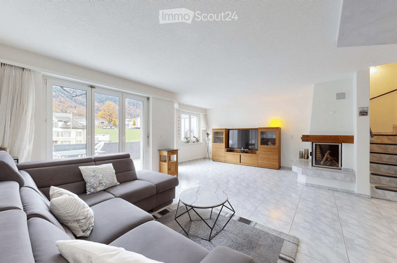 Modern living room with large windows offering a scenic view, a grey sectional sofa, a round coffee table, a fireplace, a TV cabinet, and side tables.