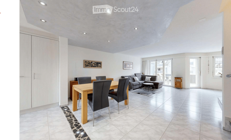 Bright living and dining area with a grey sectional sofa, wooden dining table with four black chairs, side table, and cabinet. Large windows and sliding glass doors lead to a balcony.