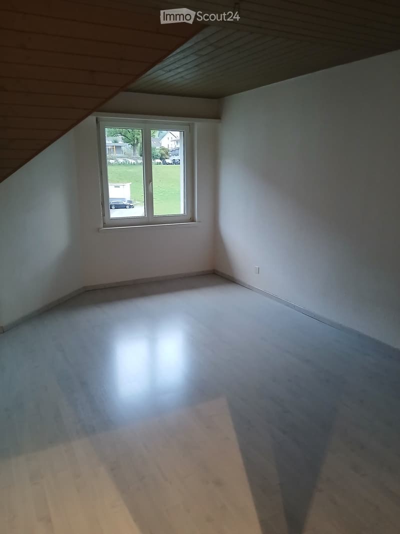 Empty room with white walls, wooden floor, and a large window with a view of the outside. The ceiling has a slanted design typical of an attic space.