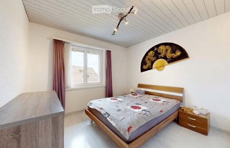 A cozy bedroom with a wooden bed, nightstand, and a decorative fan on the wall. The room features a window with curtains and a ceiling light fixture.