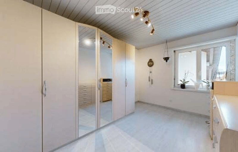 A well-lit room with a large mirrored wardrobe, a small table and chair near the window, and a potted plant on the windowsill. The room features modern lighting fixtures and a clean, minimalist design.