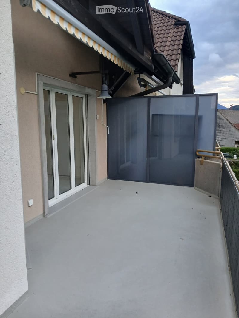 Balcony with glass railing, sliding door, and awning above, attached to a building with beige walls and a tiled roof.