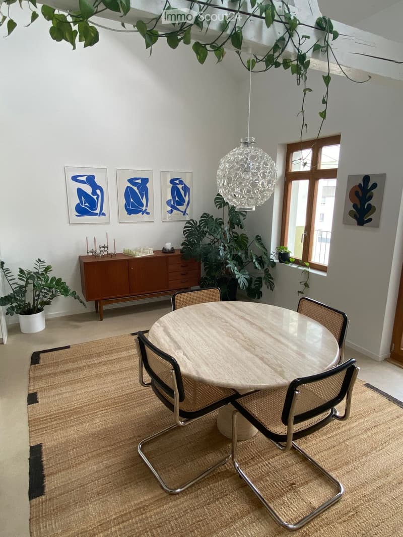 Dining area with a round wooden table, four chairs, a wooden cabinet, potted plants, artwork, and a hanging chandelier.