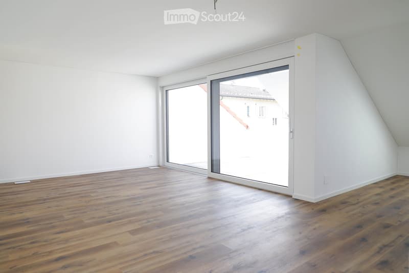 Empty attic room with wooden flooring, white walls, and large sliding glass doors leading to a view of the outside.
