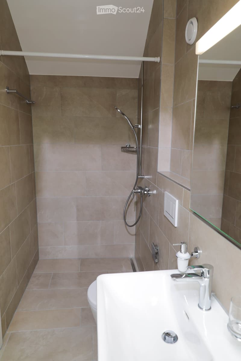 Modern bathroom with beige tiles, a white sink with a modern faucet, a toilet, a shower with a handheld sprayer, and a large mirror above the sink.