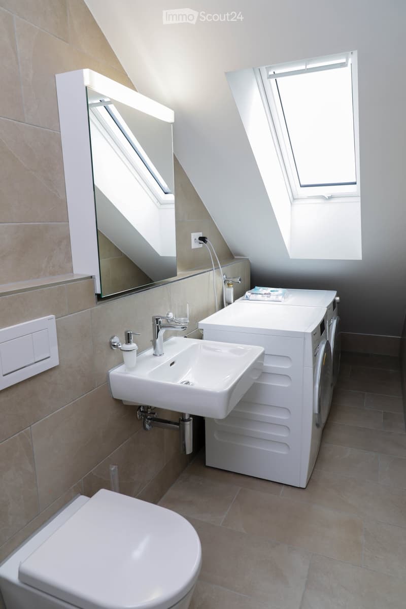 Modern bathroom with a white sink, toilet, washing machine, and large mirror. Features skylight windows and beige tiled walls.