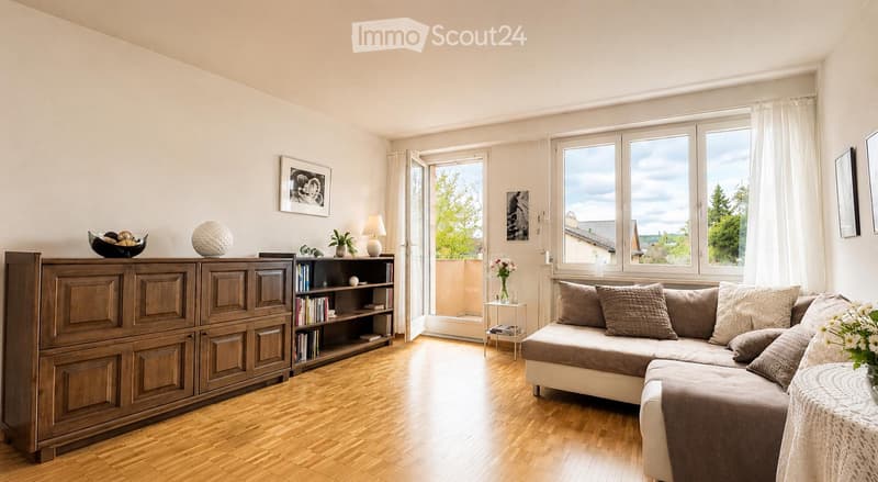 Bright living room with wooden flooring, large windows, and a balcony access. Features a sectional sofa, wooden cabinet, bookshelf, table lamp, and decorative items.