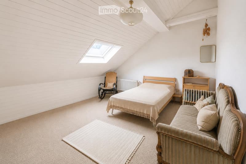 Cozy attic bedroom with a slanted ceiling, skylight window, and modern decor. Features a bed, sofa, rocking chair, and a small table.