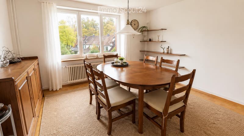 Bright dining room with a round wooden dining table surrounded by six wooden chairs with beige cushions. The room features a wooden cabinet, open shelves with decorative items, a clock, and a large window with white curtains offering a view of the street