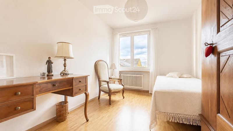 Bright bedroom with wooden flooring, a bed with white bedding, a wooden desk with a lamp and figurine, a wicker basket, and a window with a view of rooftops.
