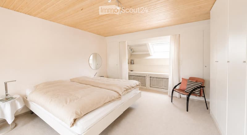 Bright, minimalist bedroom with a double bed, a small round table with a lamp, a modern chair with striped cushions, and built-in shelves with books and decor.