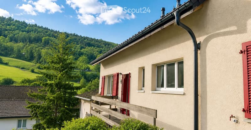 A two-story house with beige walls, red shutters, and a wooden balcony overlooking a green, hilly landscape.