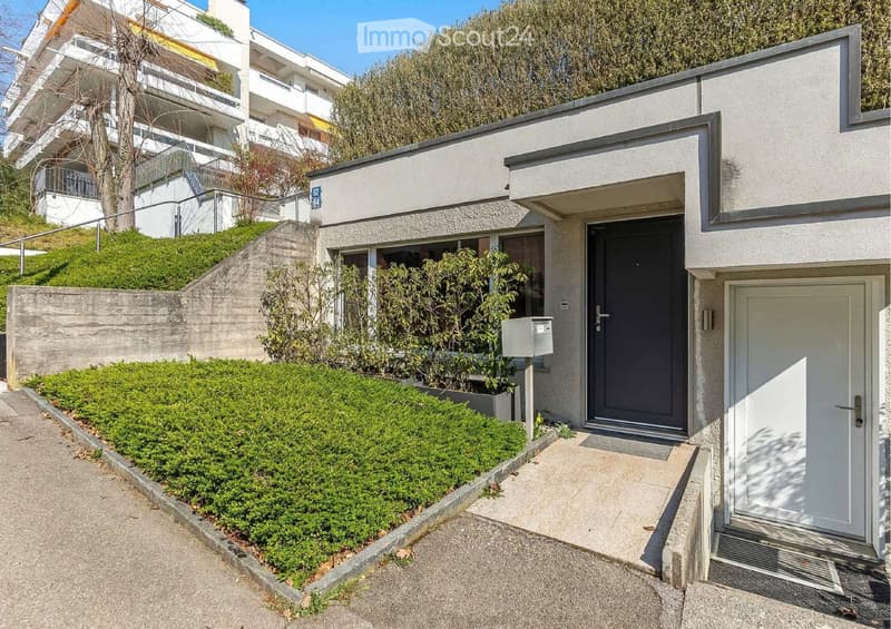 Modern two-story building with a raised ground floor, featuring a concrete facade, a small garden area with shrubs, and a mailbox near the entrance.