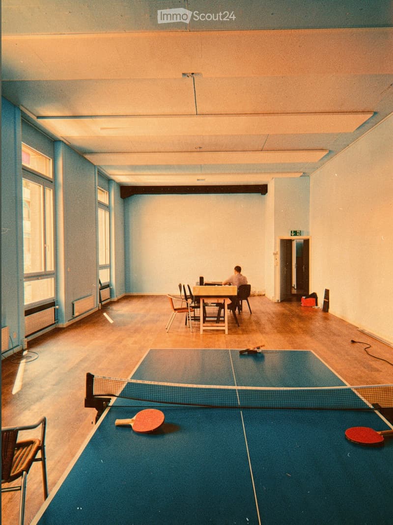 A spacious room with wooden flooring and light blue walls, featuring a ping pong table with paddles, a small table with chairs, and a person seated at the table.