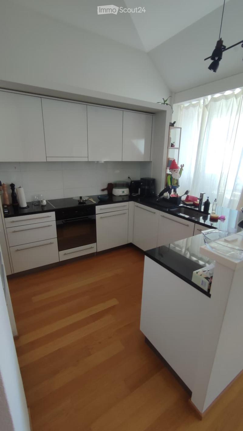 Modern kitchen with white cabinets, black countertops, and wooden flooring. Features an oven, coffee maker, sink, and various small appliances.
