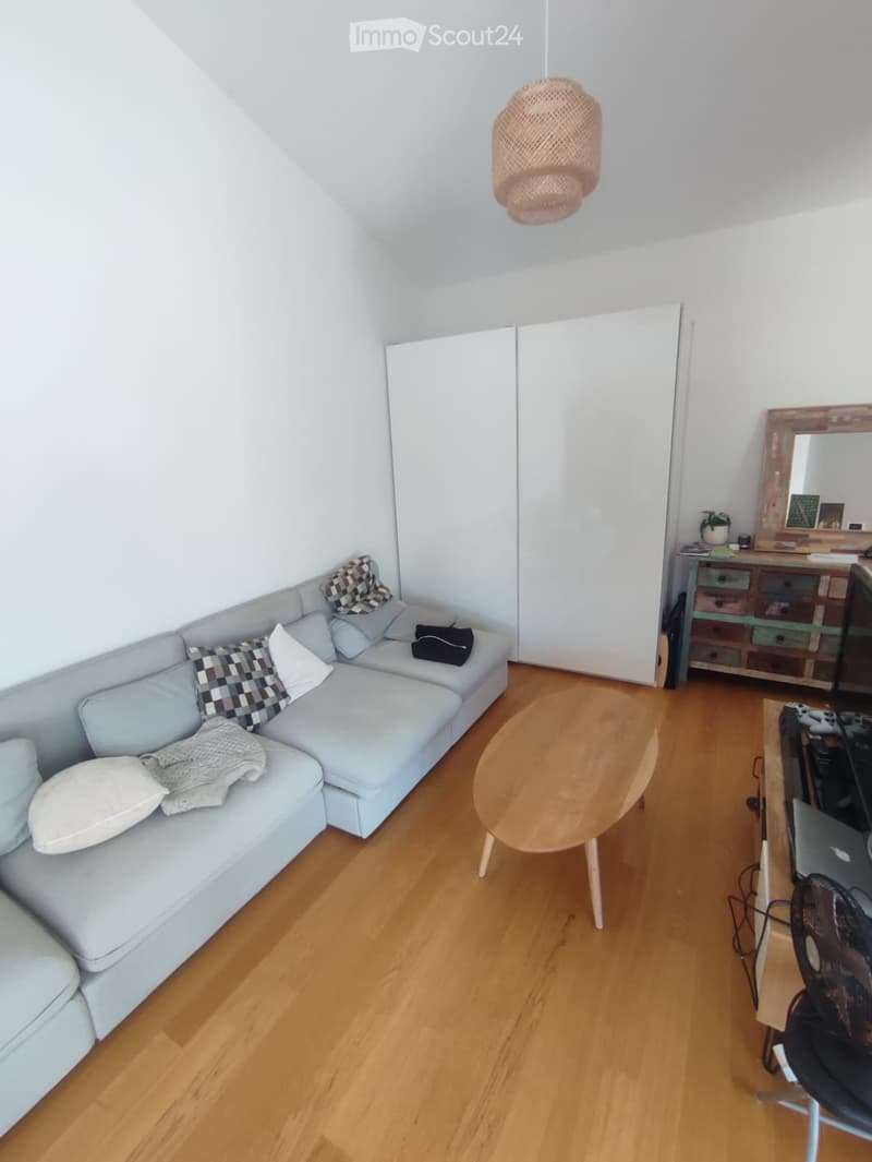 A modern living room with a light blue sectional sofa, wooden coffee table, and a small desk area with a laptop and fan. The room features white walls, wooden flooring, and a large sliding closet.