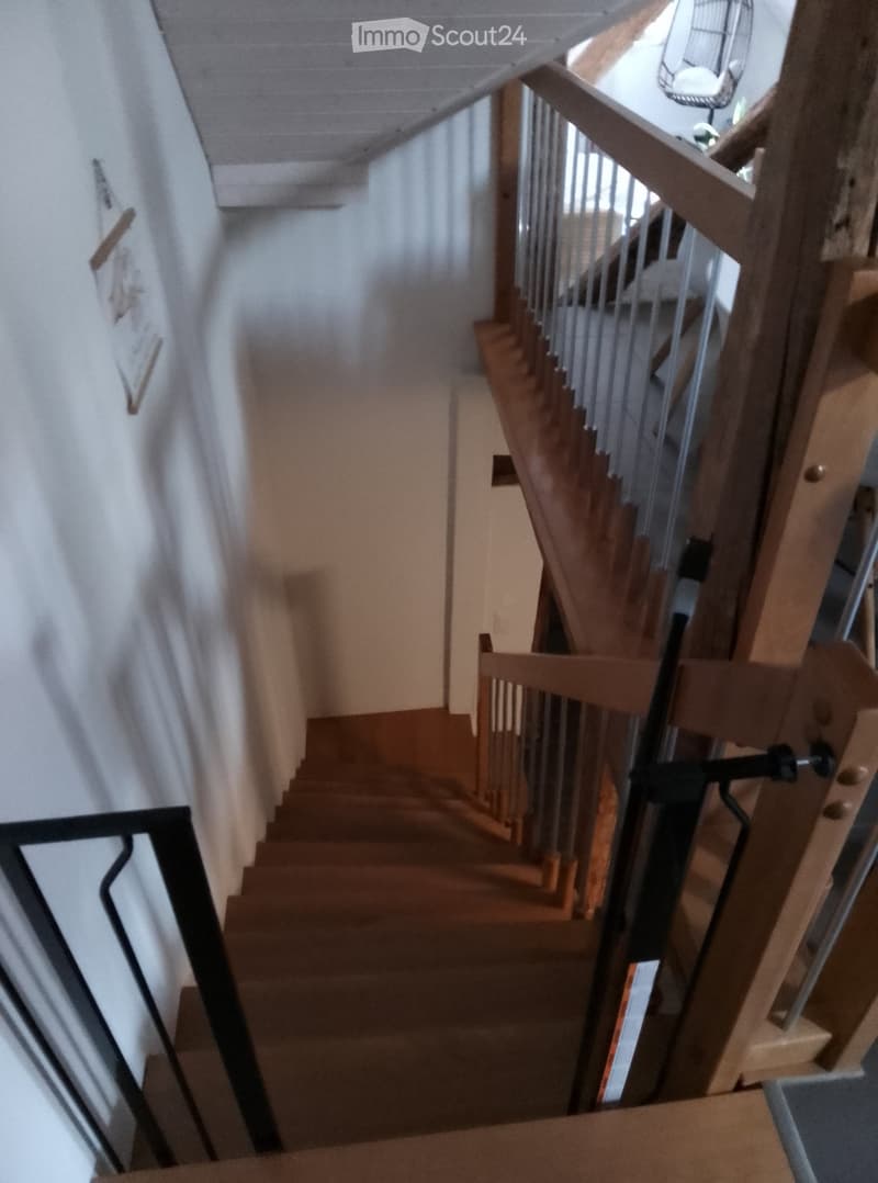 Wooden staircase with metal railing, light-colored walls, and a partially visible fan in the background.