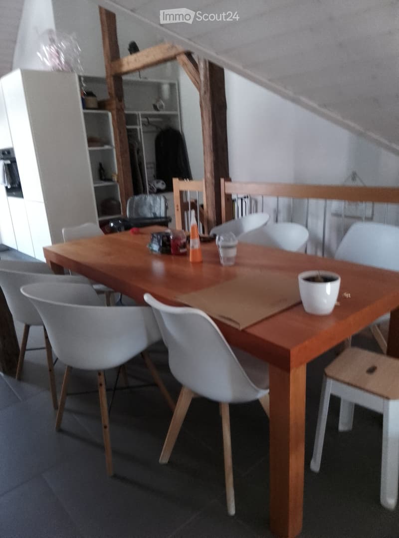 A modern dining area with a wooden table, white chairs, and a small stool. The table has a few items on it, including a cup and a small pot. The room features a wooden staircase and shelves in the background.