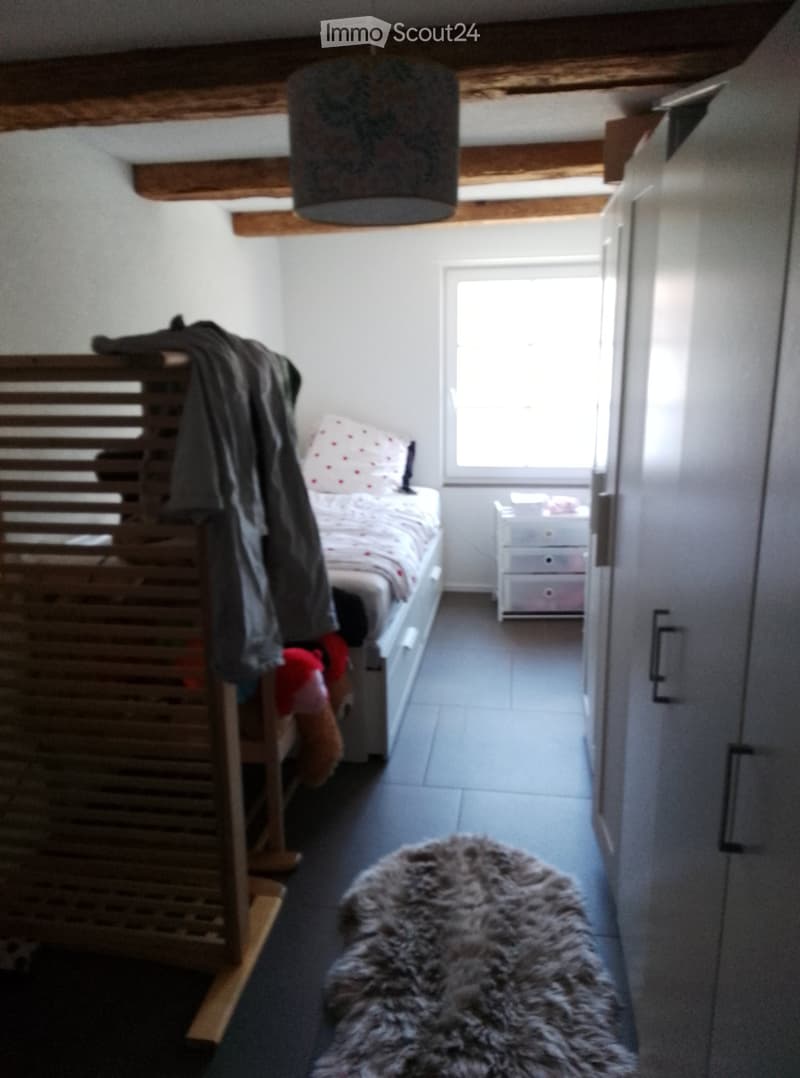 Cozy bedroom with a single bed, nightstand, fur rug, and a wooden wardrobe. Exposed wooden beams on the ceiling and a window letting in natural light.