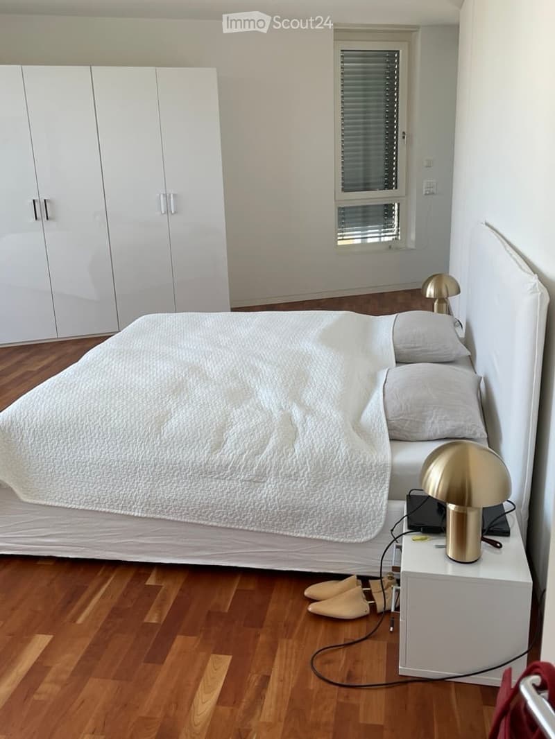 Modern bedroom with a large bed, white bedding, a nightstand with a lamp, and a white wardrobe. Wooden flooring and a window with blinds are visible.