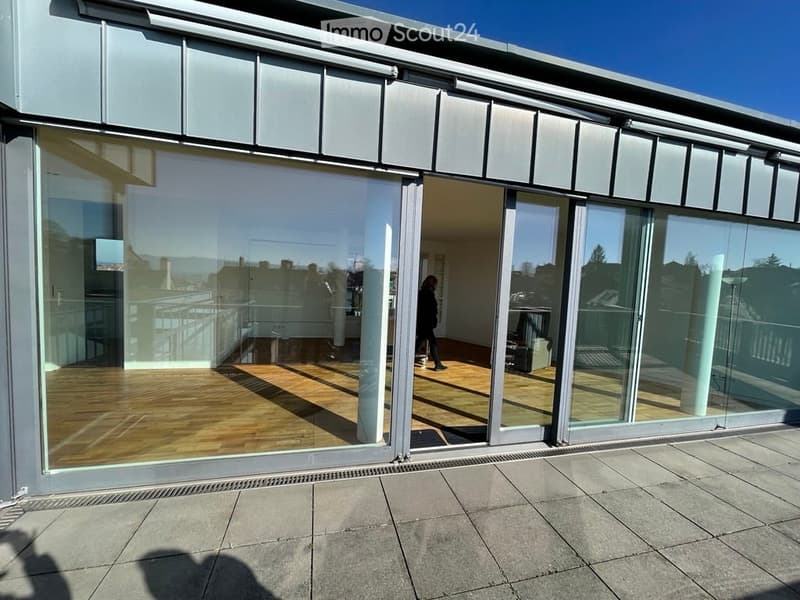 Large, modern balcony with expansive glass sliding doors, wooden flooring, and a view of the surrounding area.