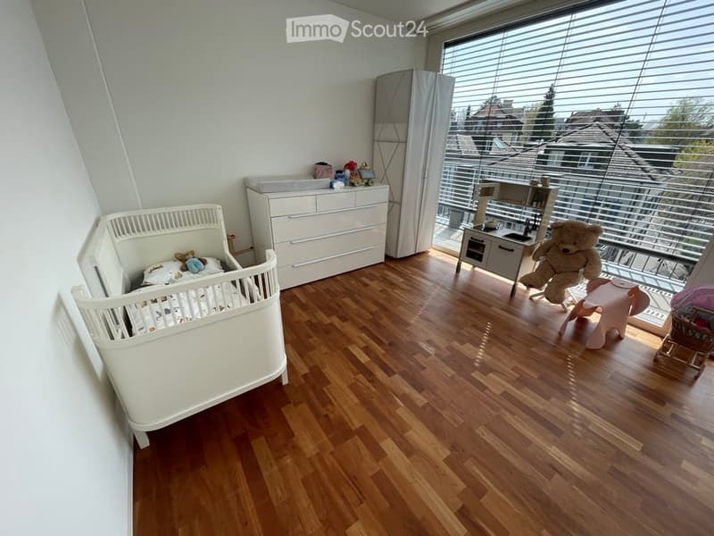 Bright, modern nursery with wooden flooring, large windows, and a balcony. Features a white crib, dresser, and various toys.