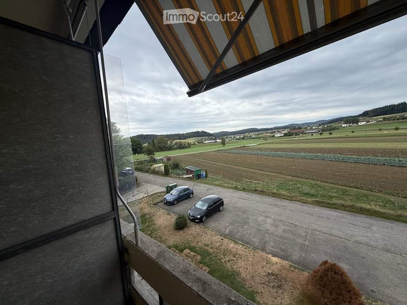 Balcony overlooking a rural landscape with parked cars, fields, and distant hills.