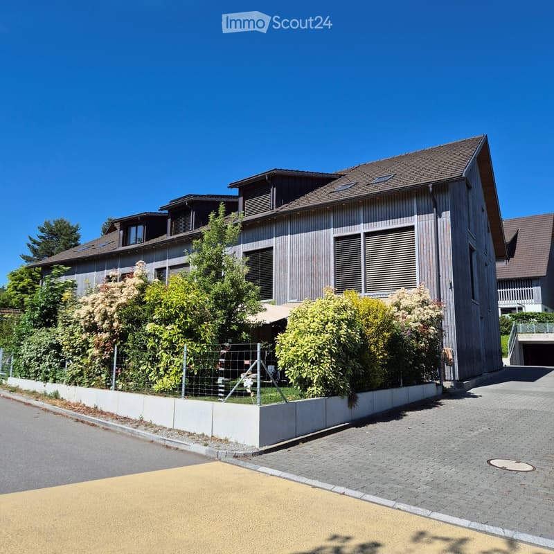 Modern two-story house with wooden exterior, multiple windows, and a fenced garden area.