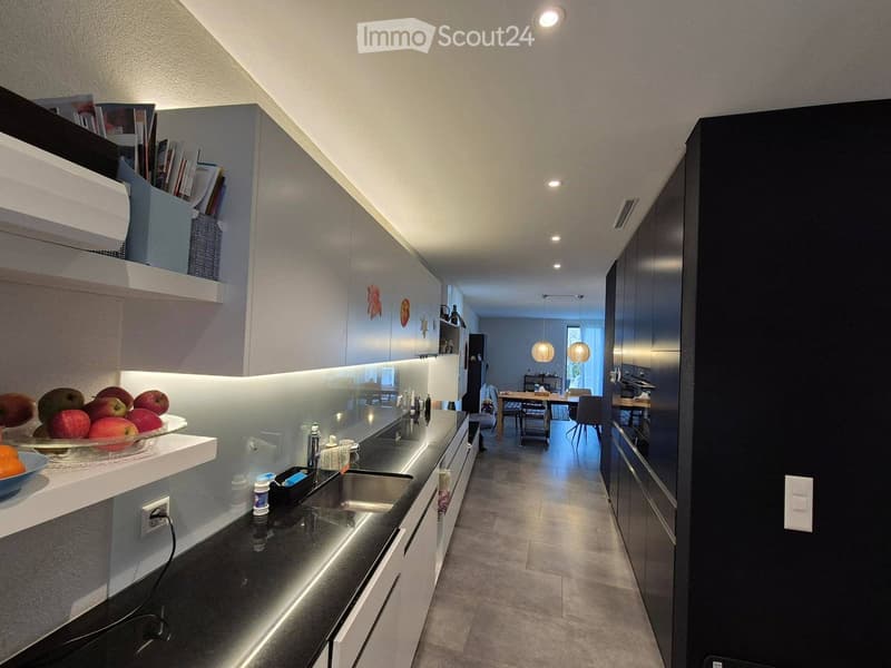 Modern kitchen with sleek black countertops, white cabinets, and built-in lighting. Visible items include a sink, faucet, and a bowl of fruit on the counter.