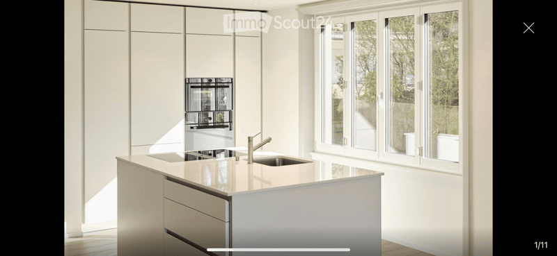 Modern kitchen with white cabinets, a central island, a double sink, and an integrated oven. Large windows provide natural light.