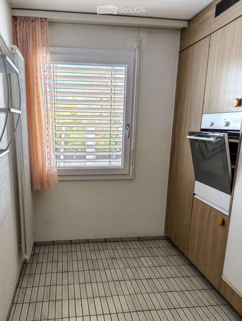 Modern kitchen with tiled floor, wooden cabinets, and a built-in oven. A window with blinds and a curtain is visible on the left side.