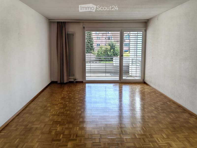 Empty room with wooden parquet flooring, white walls, large sliding glass doors leading to a balcony, and a radiator on the left side.