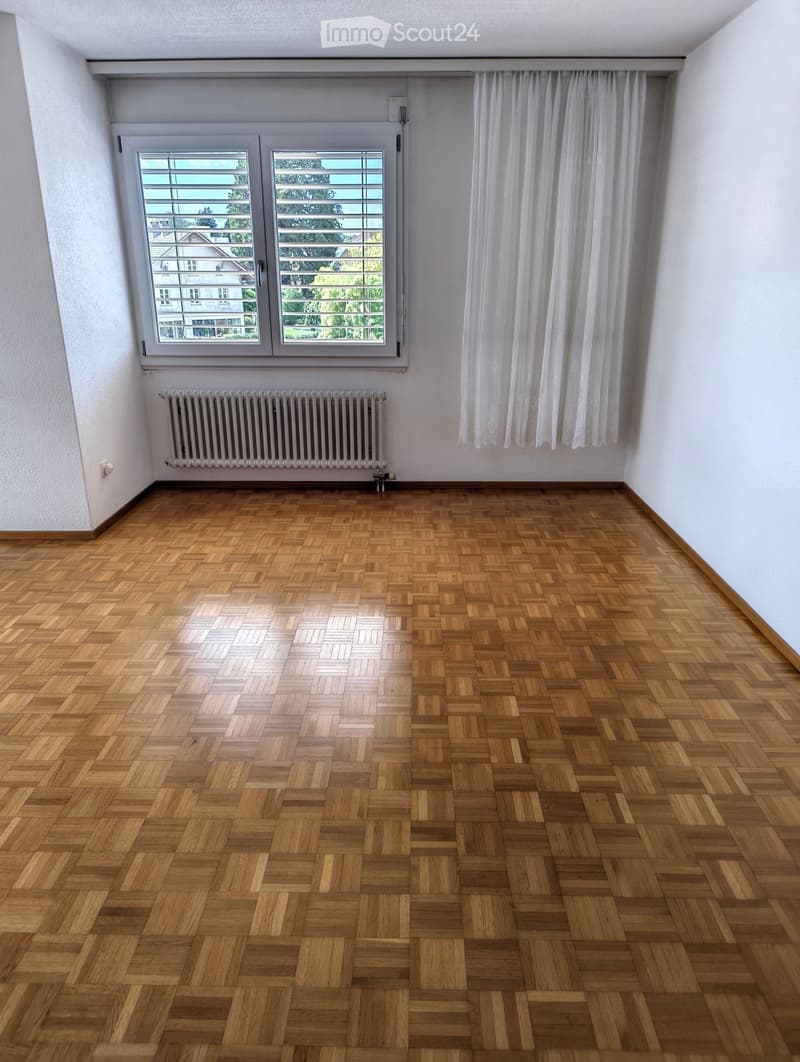 Empty room with wooden herringbone flooring, white walls, large window with white curtains, and a radiator below the window.