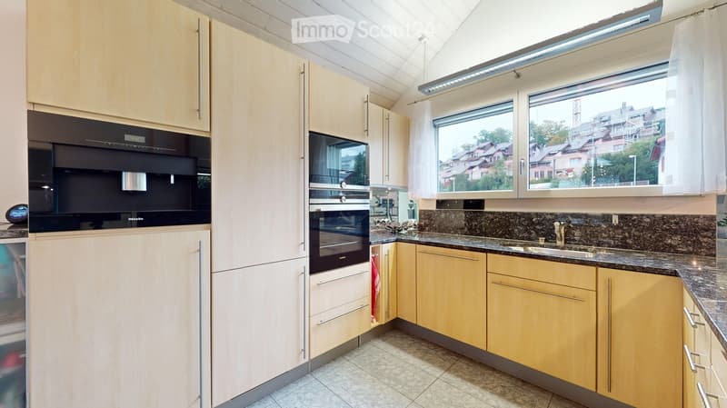 Modern kitchen with light wood cabinets, granite countertop, stainless steel appliances, and large windows overlooking a residential area.