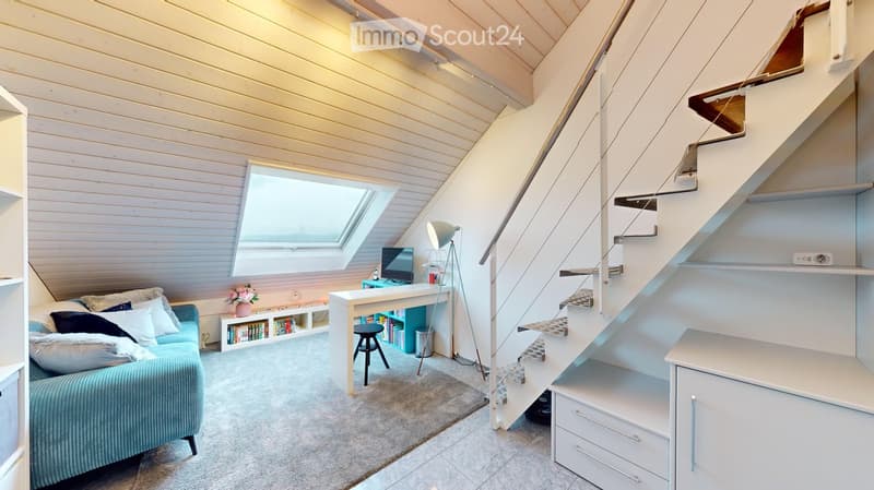 Bright, modern attic room with a slanted wooden ceiling, skylight window, and built-in staircase leading to another level. Features a blue sofa, white desk with laptop, bookshelf, and floor lamp.