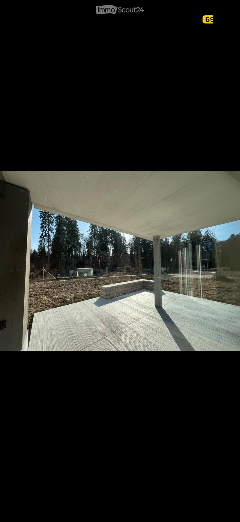 Modern outdoor patio with wooden decking, concrete bench, and large overhanging roof supported by a single column. The area is surrounded by a gravel surface and trees.
