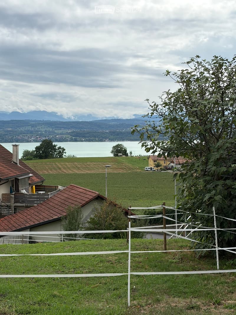 A two-story house with a red-tiled roof and a balcony, situated on a grassy hillside overlooking a lake and distant mountains.